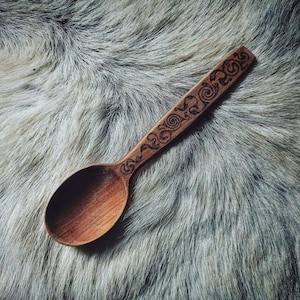 Könnte beinhalten: Ein Holzlöffel mit einem geschnitzten Griff mit einem komplizierten Spiraldesign. Der Löffel ist aus hellbraunem Holz gefertigt und liegt auf einer strukturierten, grau-weißen Pelzoberfläche. Die Löffelschale ist oval geformt.