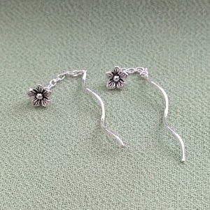 May include: A pair of silver earrings with a flower charm dangling from a chain. The earrings are made of a thin, wavy wire.