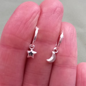 May include: A pair of silver hoop earrings. One earring has a star charm with a dark centre, and the other has a crescent moon charm. The earrings are small and delicate, suitable for everyday wear.