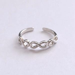 May include: A polished silver-tone ring featuring a continuous series of interconnected infinity symbols forming the band. The ring has an open design at the top, allowing for adjustment, and displays a smooth, reflective metallic finish throughout.
