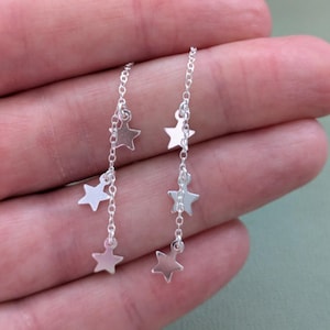 May include: Silver drop earrings with a chain design and multiple star charms. The earrings are made of a shiny, metallic material and are designed to hang down from the ear. The stars are evenly spaced along the chain.