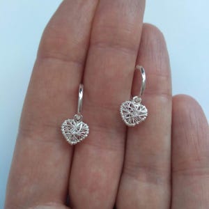 May include: A pair of silver hoop earrings featuring heart-shaped pendants. The pendants are made from fine silver wire, woven into a heart shape. The earrings are displayed against a plain backdrop.