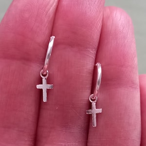 May include: A pair of silver hoop earrings with a small cross charm dangling from each hoop.