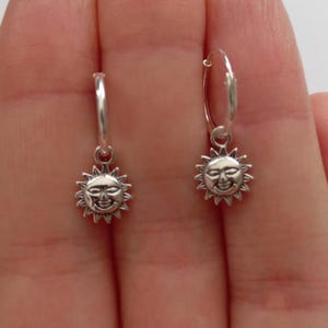 May include: A pair of silver hoop earrings featuring a smiling sun pendant. The sun charm has a detailed face and spiky rays. The earrings are displayed against a neutral background.