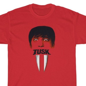May include: Red t-shirt featuring a graphic design. The design includes a face with the word "TUSK" and two white tusks. The t-shirt is made of a soft, comfortable material.