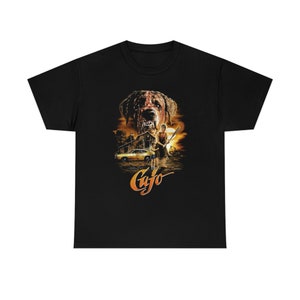 May include: Black t-shirt featuring a movie poster design. The graphic depicts a menacing dog with blood, a yellow car, and two figures. The word "Cujo" is written in orange script below the image.