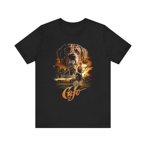 Puede incluir: Camiseta negra con un diseño de cartel de película. El gráfico representa un perro amenazante, un granero, un coche y dos personas. La palabra "Cujo" está escrita en naranja en la parte inferior del diseño.