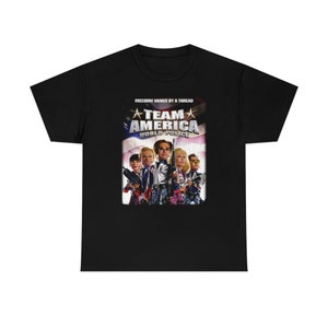 Könnte beinhalten: Schwarzes T-Shirt mit einer Grafik aus dem Film "Team America: World Police". Das Design zeigt den Filmtitel, eine Gruppe von Charakteren und den Satz "Freedom Hangs by a Thread" vor dem Hintergrund der amerikanischen Flagge.