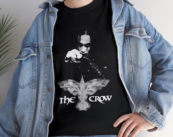 May include: Black t-shirt with a grayscale image of a person with dark makeup and a crow graphic. The text "The Crow" is below the graphic. The person is wearing a denim jacket.