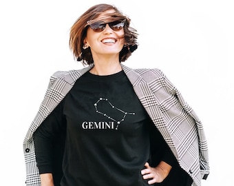 May include: A black long-sleeve shirt with the Gemini constellation graphic in white. The shirt is worn with a black and white plaid blazer and gray jeans. The person is wearing sunglasses and smiling.