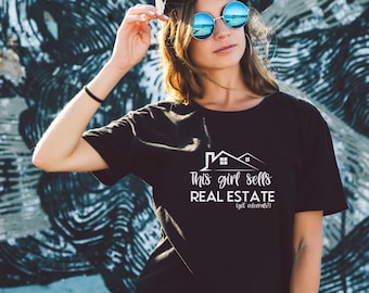 May include: Black t-shirt with white text that reads "This girl sells REAL ESTATE (got referrals?)". The shirt features a graphic of two houses. The person is wearing sunglasses and a black hat.