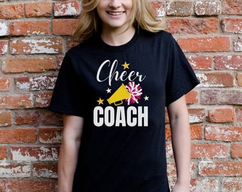 Peut inclure: T-shirt noir avec l'inscription blanche "Cheer COACH", un mégaphone jaune et un motif pompon rose et blanc. Le mot "Cheer" est en cursive, et il y a des étoiles dorées.