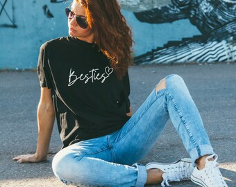 Puede incluir: Camiseta negra con la palabra "Besties" en escritura blanca y un corazón. La camiseta se combina con vaqueros rotos de color azul claro y zapatillas blancas. La persona está sentada en una superficie de hormigón con un fondo azul y gris.