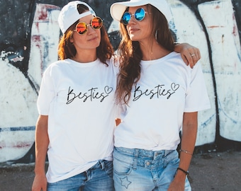 Puede incluir: Dos personas con camisetas blancas con la palabra "Besties" en escritura negra y un símbolo de corazón. También llevan gorras de béisbol blancas y gafas de sol redondas con lentes reflectantes. Llevan vaqueros azules.