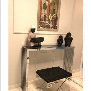 May include: A silver metal console table with a black velvet cushioned stool. The table has a mirrored surface and a decorative design. There are two black ceramic vases with gold accents on the table.