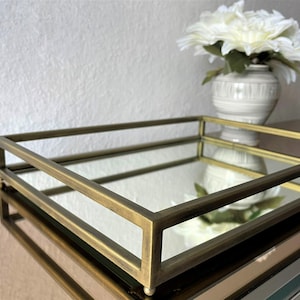 Puede incluir: Una bandeja rectangular dorada con una superficie de espejo. La bandeja tiene un borde elevado y es perfecta para exhibir artículos decorativos o servir aperitivos.