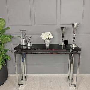 May include: A contemporary console table featuring a black glass top and a chrome metal frame. It's adorned with white flowers in a silver vase, two silver vases, and two geometric candle holders. The backdrop is a grey wall.