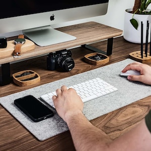 Peut inclure: Un organisateur de bureau en bois avec un support d'écran, un chargeur sans fil, un support de montre, un appareil photo et un casque. L'organisateur de bureau est sur un bureau en bois avec un tapis de souris en feutre gris et un clavier blanc.