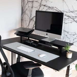 May include: A black standing desk with a black monitor stand, a laptop stand, a keyboard, a mouse, and a potted plant. The desk is in front of a wall with a white and gray patterned wallpaper.