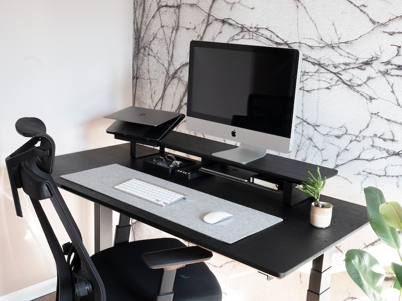 May include: A black standing desk with a black monitor stand, a laptop stand, a keyboard, a mouse, and a potted plant. The desk is in front of a wall with a white and gray patterned wallpaper.