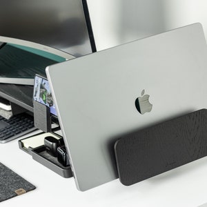 May include: A silver laptop computer with an Apple logo on the back is propped up on a black wooden stand. The laptop is on a white desk with a black keyboard and a black phone charger.