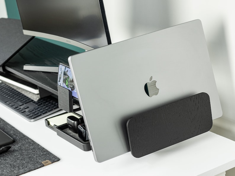 May include: A silver laptop computer with an Apple logo on the back is propped up on a black wooden stand. The laptop is on a white desk with a black keyboard and a black phone charger.