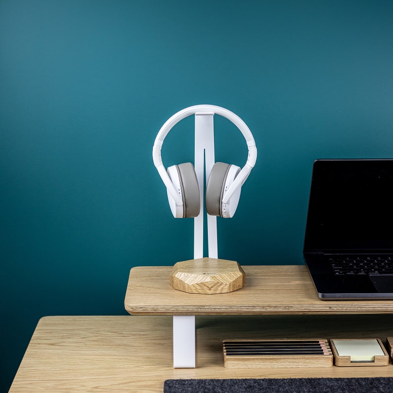 Puede incluir: Soporte para auriculares blanco con base de madera y un port&aacute;til en un escritorio de madera. Los auriculares son grises y blancos.