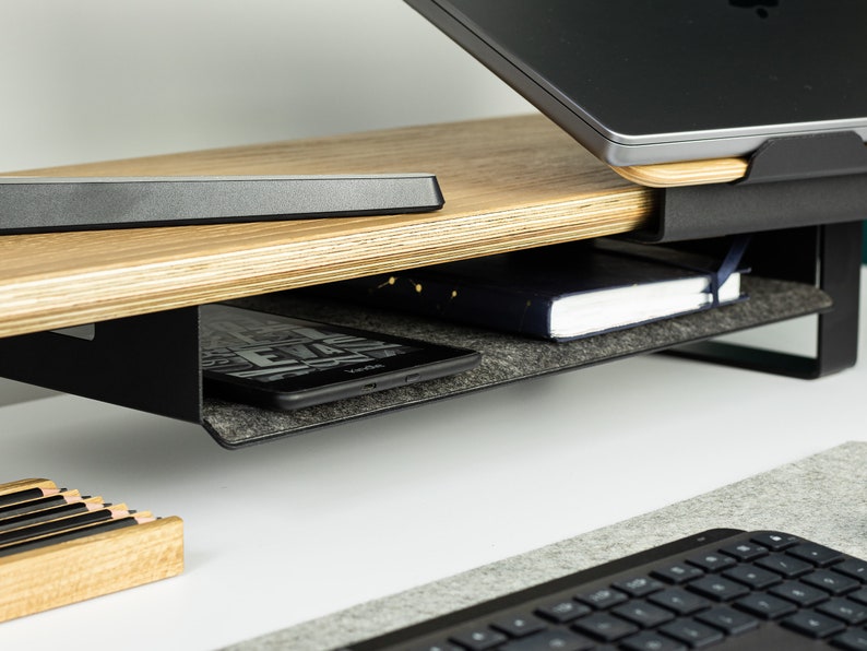 May include: A black metal laptop stand with a wooden top and a gray felt-lined drawer. The stand has a built-in shelf for storing items like a book or a tablet.