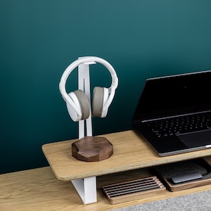 Puede incluir: Soporte para auriculares blanco con base de madera y un par de auriculares blancos en el soporte. Un ordenador port&aacute;til est&aacute; en un estante de madera debajo del soporte.