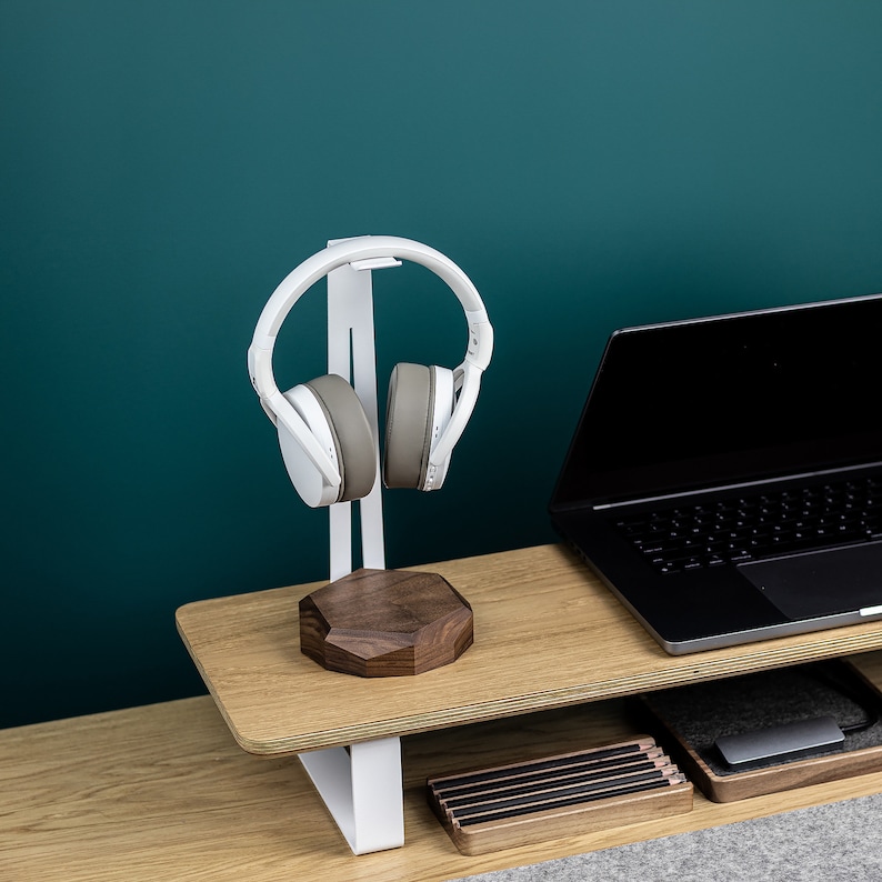 Puede incluir: Soporte para auriculares blanco con base de madera y un par de auriculares blancos en el soporte. Un ordenador port&aacute;til est&aacute; en un estante de madera debajo del soporte.