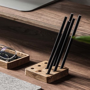 May include: A wooden pencil holder with a grid of holes to hold pencils. The holder is made of light brown wood and has a natural finish. Six black pencils are standing upright in the holder.
