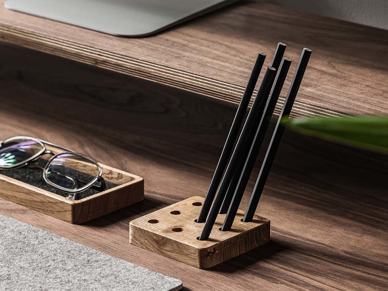 May include: A wooden pencil holder with a grid of holes to hold pencils. The holder is made of light brown wood and has a natural finish. Six black pencils are standing upright in the holder.
