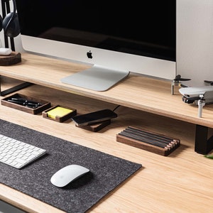 Pu&ograve; includere: Un supporto per monitor in legno con una cornice in metallo nero, un tappetino per mouse grigio, una tastiera bianca, un mouse grigio e un piccolo drone sulla scrivania. Ci sono anche diversi piccoli organizzatori in legno sulla scrivania.