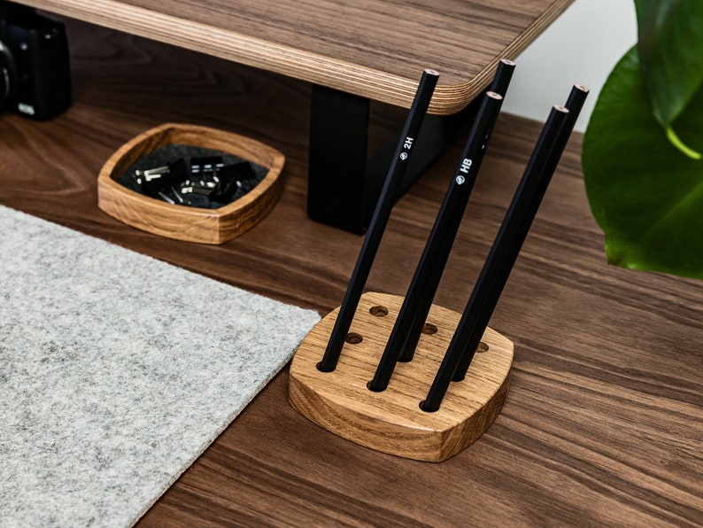 May include: A wooden pencil holder with six slots, holding five black pencils. The pencils are labelled with the grades "2H" and "HB".