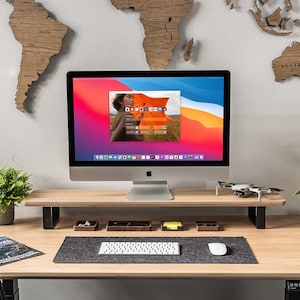 Pu&ograve; includere: Un ambiente di lavoro moderno con una scrivania in legno, un tappetino per mouse nero e grigio, una tastiera bianca, un drone bianco, una pianta in vaso e un iMac argentato con uno schermo che mostra uno sfondo desktop con una donna in piedi davanti a un design astratto colorato.