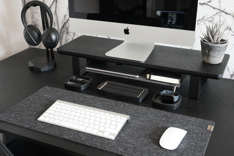 Pu&ograve; includere: Una scrivania nera con un supporto per monitor nero, una tastiera bianca, un mouse bianco e un supporto per cuffie nero con cuffie nere. La scrivania ha un tappetino per mouse in feltro grigio.