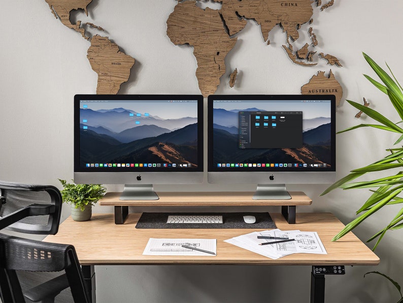 Puede incluir: Dos ordenadores iMac plateados con pantallas negras que muestran un icono de carpeta en la pantalla de la derecha y una cordillera en la pantalla de la izquierda. Los ordenadores est&aacute;n sobre un escritorio de madera con un teclado negro, un rat&oacute;n y una planta en una maceta blanca. Un mapa del mundo de madera est&aacute; en la pared detr&aacute;s de los ordenadores.