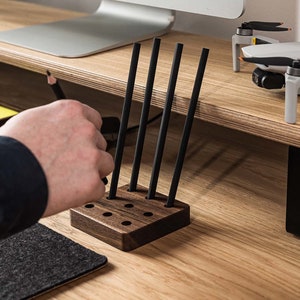 May include: A wooden pencil holder with five slots for pencils. The holder is made of dark wood and has a natural finish. Five black pencils are in the holder.