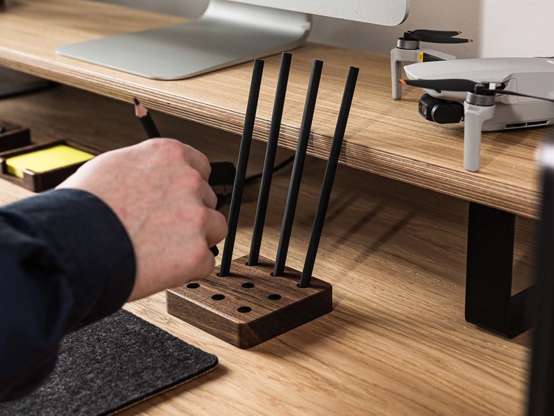 May include: A wooden pencil holder with five slots for pencils. The holder is made of dark wood and has a natural finish. Five black pencils are in the holder.