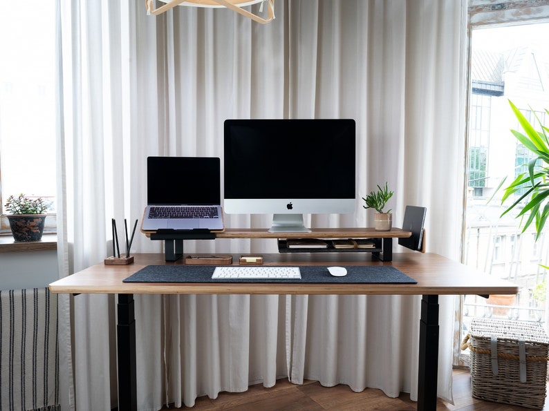 May include: A standing desk with a black frame and a light wood top. A laptop and a desktop computer are on a wooden stand on the desk. A white keyboard and a white mouse are on the desk. A small potted plant is on the stand.