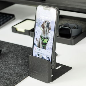 May include: A black wooden phone stand with a smartphone in a black case. The phone screen shows a photo of a dog in the snow. The time on the phone screen is 16:00.