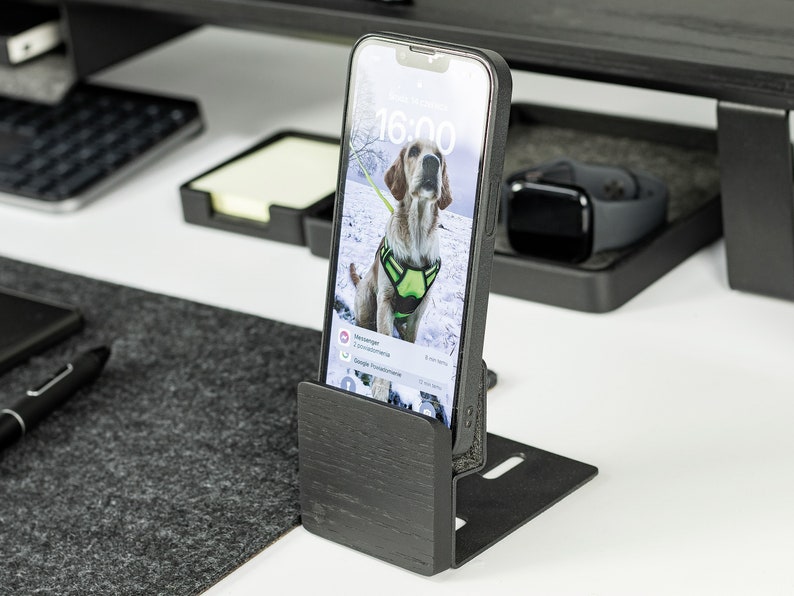 May include: A black wooden phone stand with a smartphone in a black case. The phone screen shows a photo of a dog in the snow. The time on the phone screen is 16:00.
