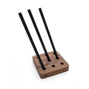 May include: A wooden stand with nine holes, designed to hold three black pens. The stand is made of dark wood and has a square shape.