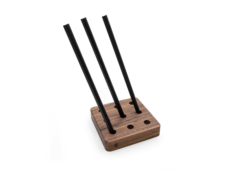 May include: A wooden stand with nine holes, designed to hold three black pens. The stand is made of dark wood and has a square shape.