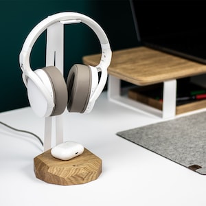 Puede incluir: Auriculares blancos con una almohadilla gris en un soporte blanco con una base de madera. Una funda de carga inal&aacute;mbrica blanca est&aacute; en la base.