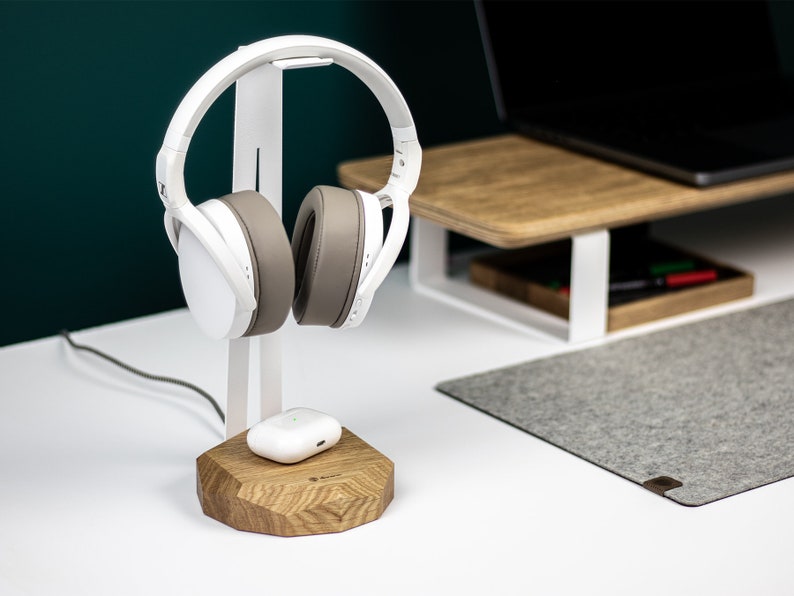 Puede incluir: Auriculares blancos con una almohadilla gris en un soporte blanco con una base de madera. Una funda de carga inal&aacute;mbrica blanca est&aacute; en la base.