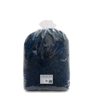 Op de afbeelding: Een grote, doorzichtige plastic zak gevuld met donkerblauwe gerimpelde papieren stroken. De zak heeft een label met de tekst "Crinkle Paper Shreds Eco-friendly & multi-purpose 1kg Dark Blue".