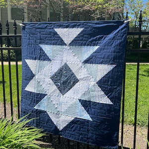 Könnte beinhalten: Eine marineblaue Steppdecke mit einem großen weißen und hellblauen Sternmuster. Der Stern ist auf der Steppdecke zentriert und hat ein dunkelblaues Quadrat in der Mitte. Die Steppdecke hängt an einem schwarzen Metallzaun.