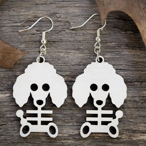 May include: A pair of white wooden earrings shaped like poodles with a skeleton design. The earrings are hanging from silver hooks.