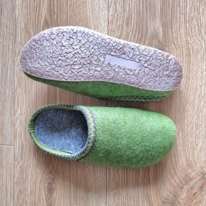 Puede incluir: Un par de zapatillas de fieltro verde con interior gris y suela marr&oacute;n texturizada. Las zapatillas tienen un ribete verde oscuro y gris alrededor de la abertura. Las zapatillas se muestran sobre una superficie de madera marr&oacute;n claro.